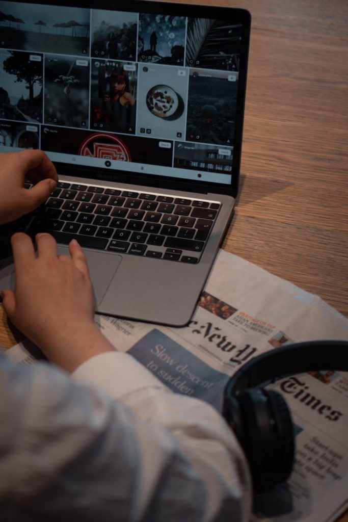 A person working on a laptop placed on a wooden desk with a newspaper and headphones nearby.